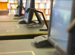 Computer monitors on library desks with bookshelves in the background.